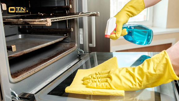  A person wearing yellow gloves is cleaning a dirty oven door with a blue spray bottle and a yellow cloth, focusing on removing grime from the glass.