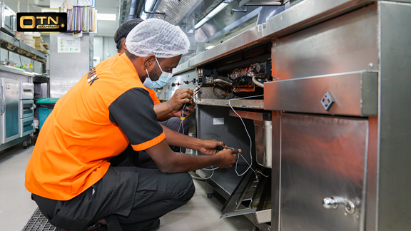 A technician in an orange shirt and hairnet repairs and maintenance commercial kitchen machinery with tools in a kitchen setting