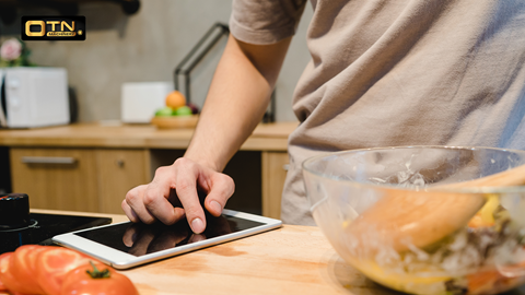 Person using a tablet in the kitchen while preparing food, with tomatoes and a salad bowl in the foreground.