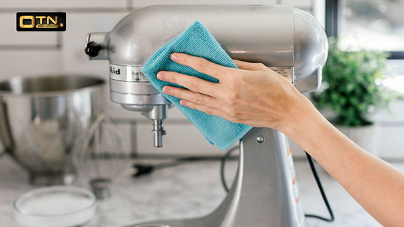 A person's hand wiping a silver kitchenaid stand mixer with a blue cloth in a bright kitchen.