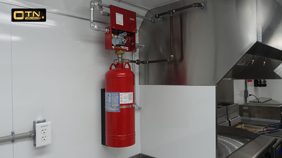 A fire suppression system is installed in a commercial kitchen to meet fire safety regulations. A red fire extinguisher cylinder is attached to a wall, with connected piping and an exhaust hood nearby.