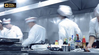 Chefs dressed in white uniforms and hats are working busily in a commercial kitchen with various ingredients and utensils on the countertop, all while adhering to strict kitchen safety protocols.