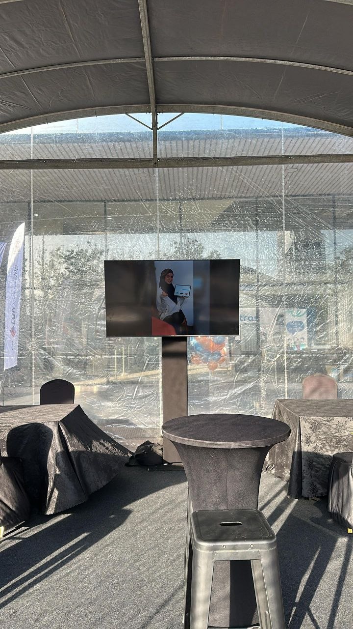 A television mounted on a pedestal stand displays a woman holding a tablet inside a clear-walled roadshow tent.