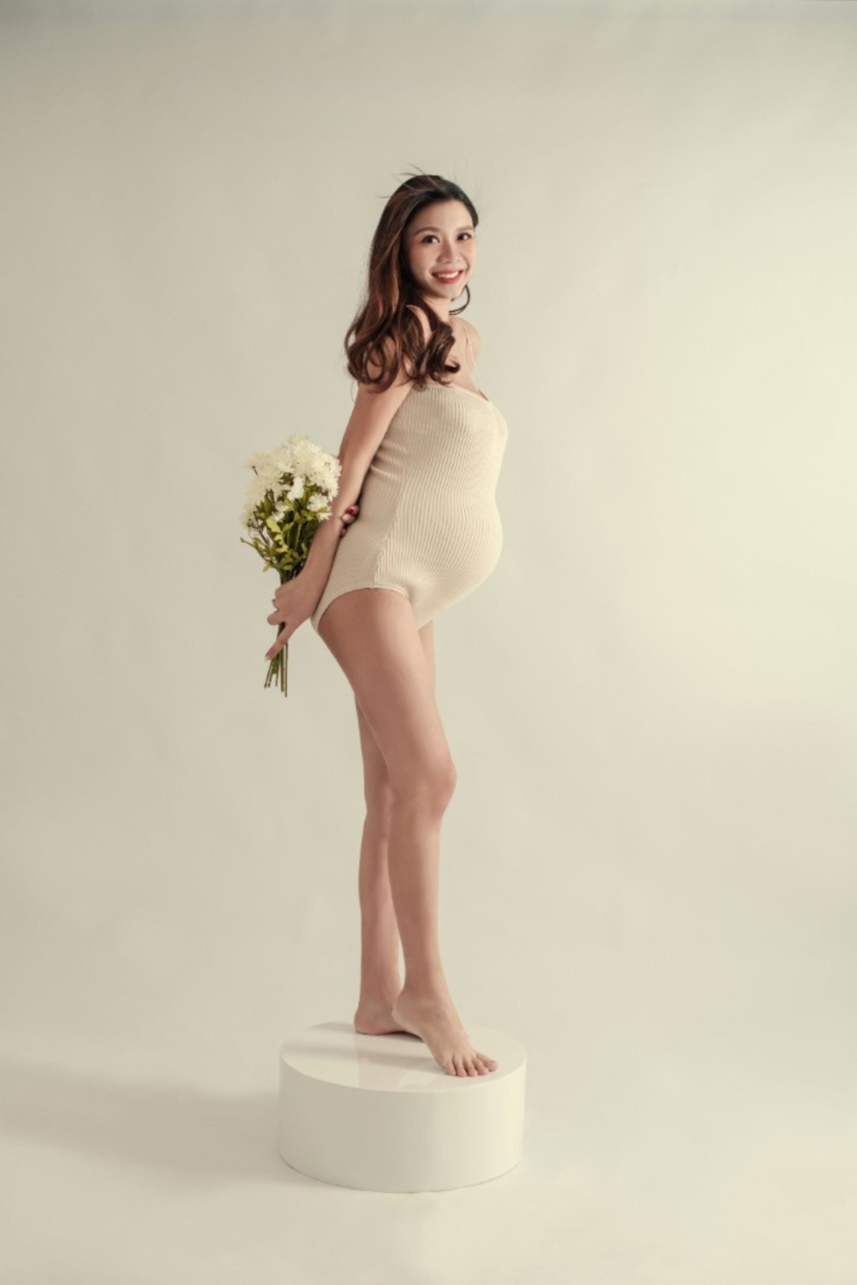 Elegant maternity photography portrait of expecting mother standing with flowers in a minimal studio